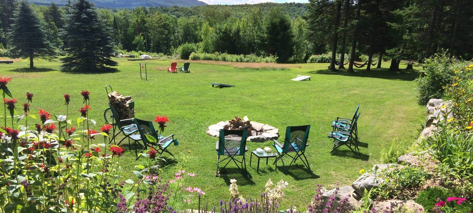 Grassy common area with yard games, chairs, and campfire surrounded by flowers, pine trees and distant hills