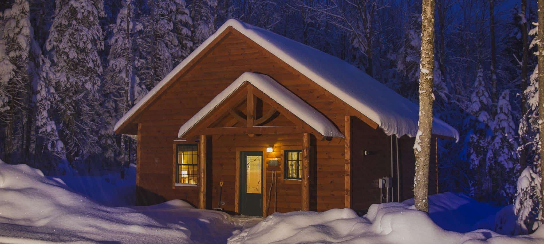 Log cabin with steep gable roof covered in snow at dusk surrounded by snow-covered trees and ground