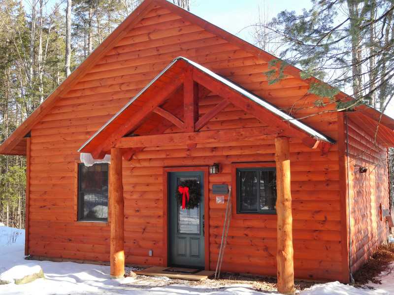 A warm, sunlit timber-frame cabin with reddish-brown wood siding and a gabled porch, nestled among tall pine trees at Robert Frost Mountain Cabins in Ripton, Vermont.