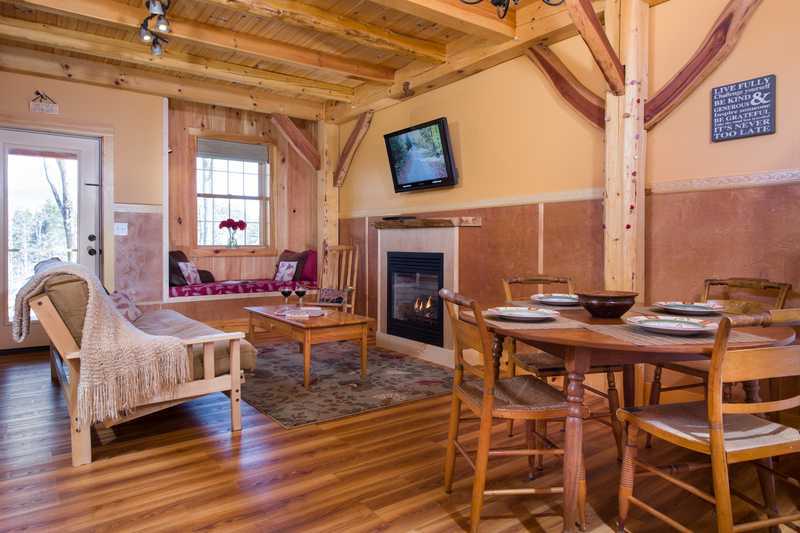 A spacious timber-frame cabin interior featuring an open-concept living and dining area with a round wooden table, a cozy fireplace, and a built-in Swedish reading nook by a window. The room is characterized by warm wood tones, exposed structural beams, and large windows that fill the space with natural light.