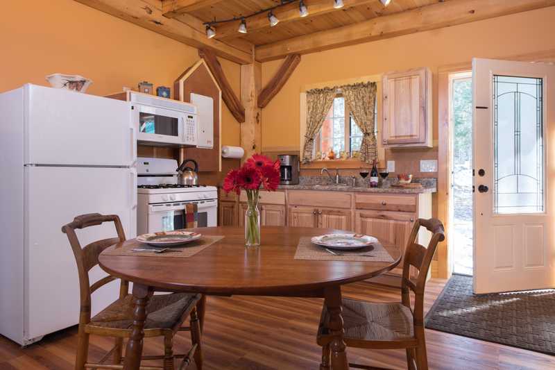 A bright, open-concept cabin kitchen and dining area with an oval wooden table and chairs in the foreground. The space features light wood cabinetry, white appliances, and warm orange walls, all framed by rustic timber-frame beams and natural light from an open door.