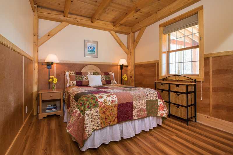 An inviting cabin bedroom featuring a queen-size bed with a colorful patchwork quilt, framed by rustic timber beams and warm, wood-paneled walls. A large window provides natural light, complemented by bedside lamps, a wooden nightstand with yellow flowers, and a matching dresser.