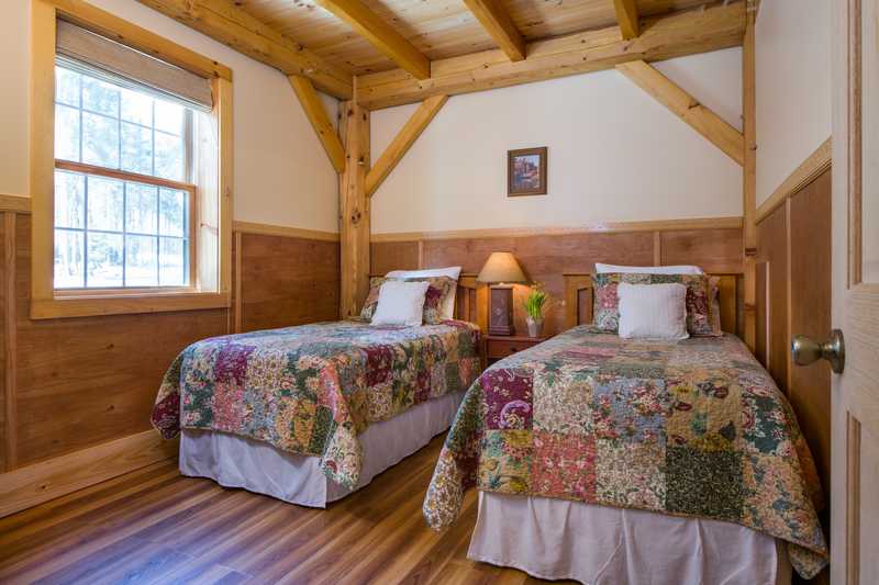 A bright cabin bedroom featuring two twin beds with vibrant, multi-colored floral patchwork quilts. The room is constructed with light-colored timber-frame beams and natural wood wainscoting, with a large window on the left letting in plenty of daylight.