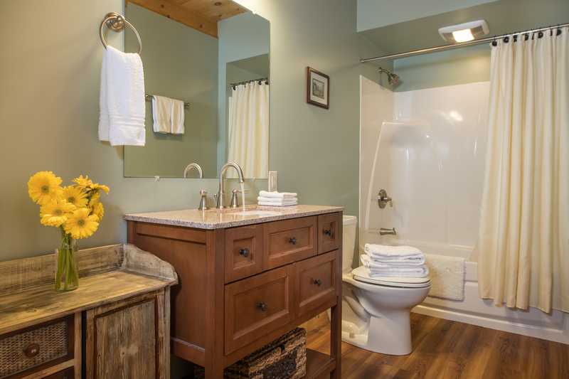 A clean, updated cabin bathroom featuring a warm wood vanity with a white countertop and a large mirror. The room is decorated with a vase of bright yellow sunflowers, neatly stacked white towels, and a simple cream-colored shower curtain, creating a bright and welcoming atmosphere.