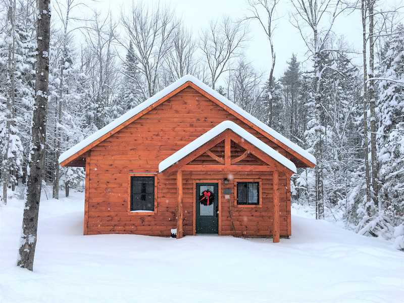 A small, wood-sided cabin with a peaked roof and a timber-frame porch entryway in a dense forest during winter. The cabin's warm, orange-toned wood stands out against the white snow covering the ground and the surrounding trees under a bright sky.