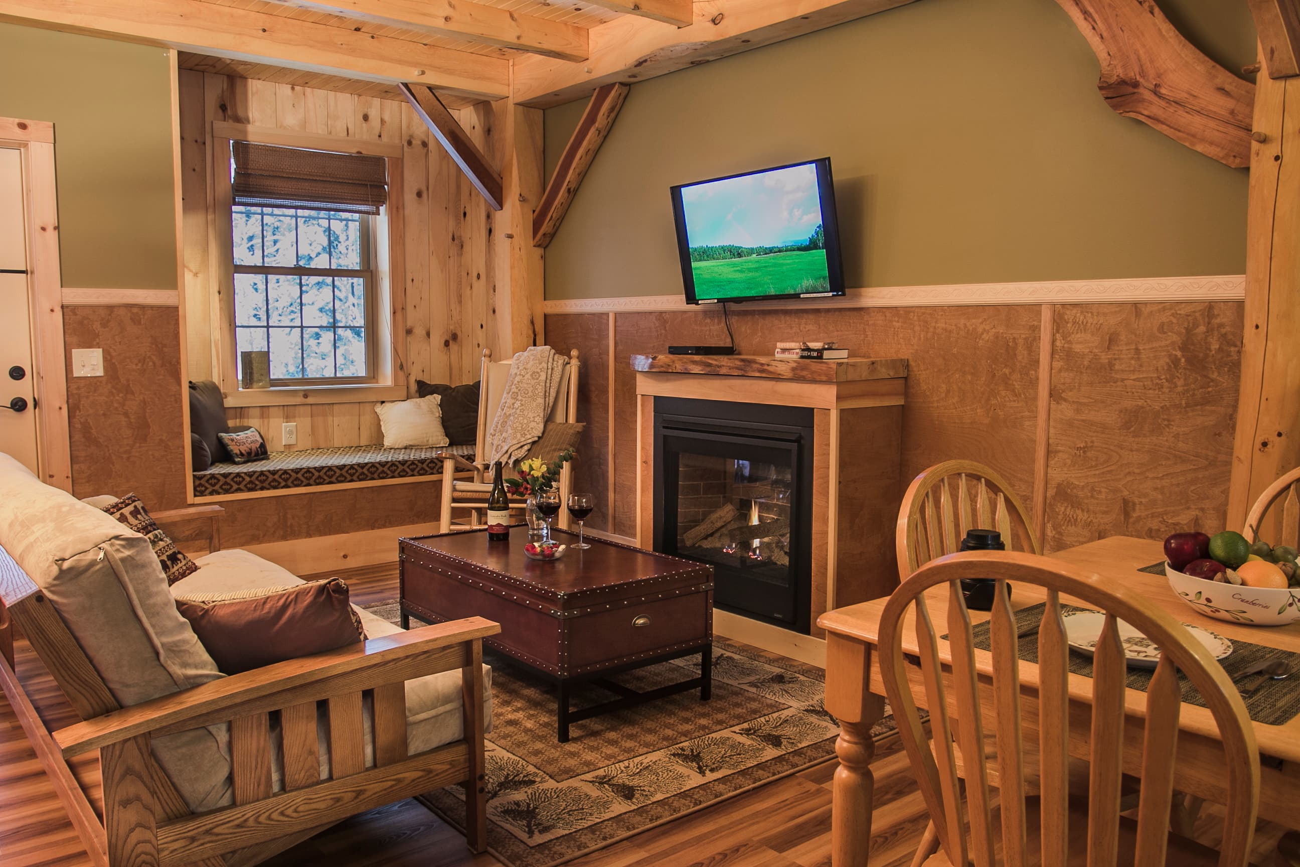 A cozy living room inside a rustic cabin features a wood-frame sofa with light cushions, a dark wood coffee table on a patterned rug, and a small dining table with wooden chairs in the foreground. The room is decorated with sage green walls, wood-paneled wainscoting, and exposed timber beams, and includes a fireplace with a TV mounted above it and a built-in window seat in the background.