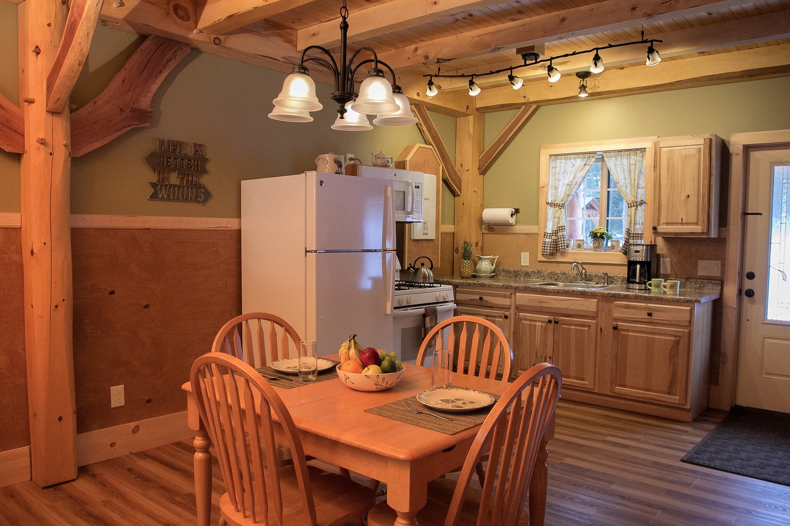 The interior of a rustic cabin features a kitchen and dining area with sage green walls, wood wainscoting, and exposed timber beams. A wooden dining table with four matching chairs is set in the foreground. The kitchen in the background includes light wood cabinetry, a white refrigerator, and a window with white curtains, complemented by warm wood flooring.