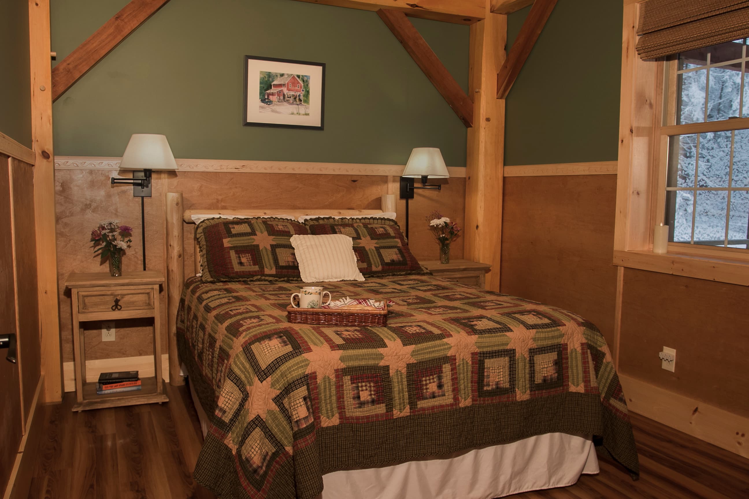 A cozy bedroom in a rustic cabin features a bed with a log-style frame and an earth-toned geometric patchwork quilt. The room is decorated with sage green walls, wood-paneled wainscoting, and exposed timber beams, with a window on the right and two wooden nightstands with lamps flanking the bed.