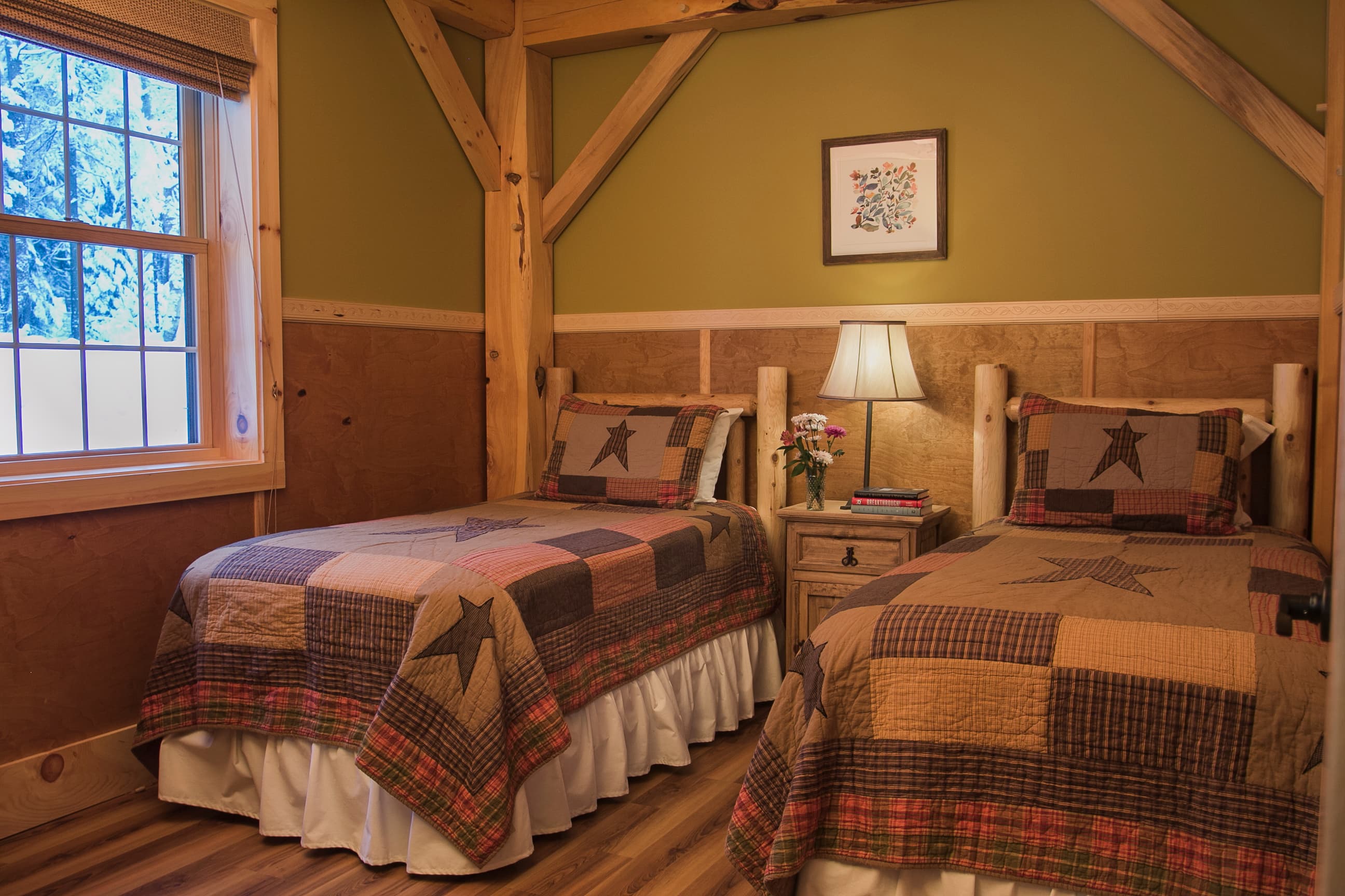 A bedroom in a rustic cabin featuring two twin beds with log-style frames and earth-toned patchwork quilts with star patterns. The room has olive green walls, wood-paneled wainscoting, and exposed timber beams, with a window on the left and a small wooden nightstand with a lamp positioned between the beds.