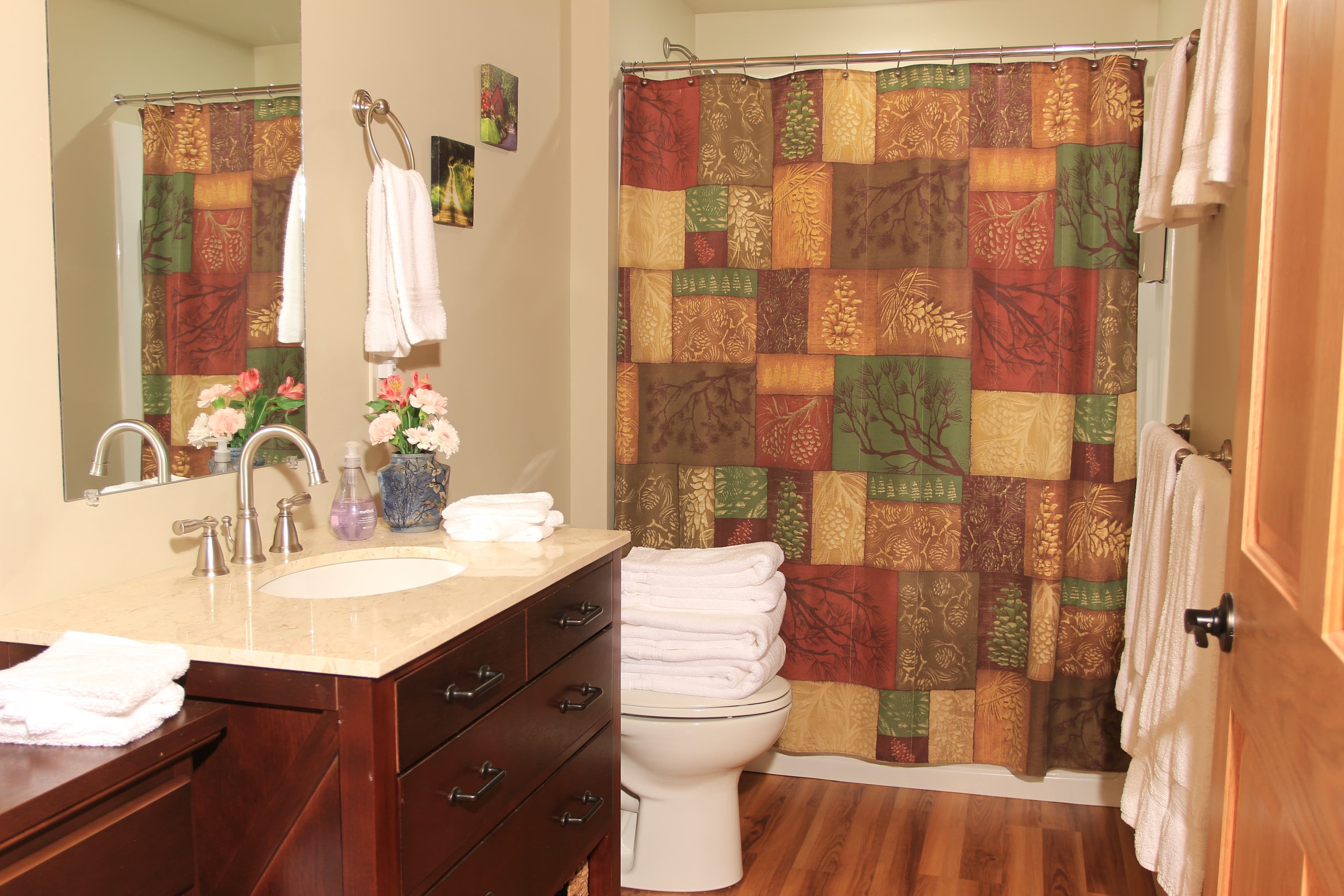 A bathroom inside a rustic cabin featuring a dark wood vanity with a white sink and a large mirror. The room has light green walls, wood flooring, and a shower-tub combination with a colorful, multi-patterned patchwork shower curtain.
