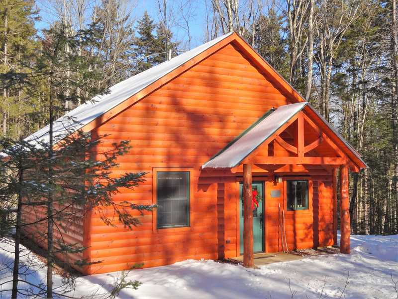 A small, wood-sided cabin with a peaked roof and a timber-frame porch entryway stands in a snowy forest. The bright orange-toned wood of the cabin contrasts with the white snow on the ground and the surrounding bare and evergreen trees under a clear blue sky.