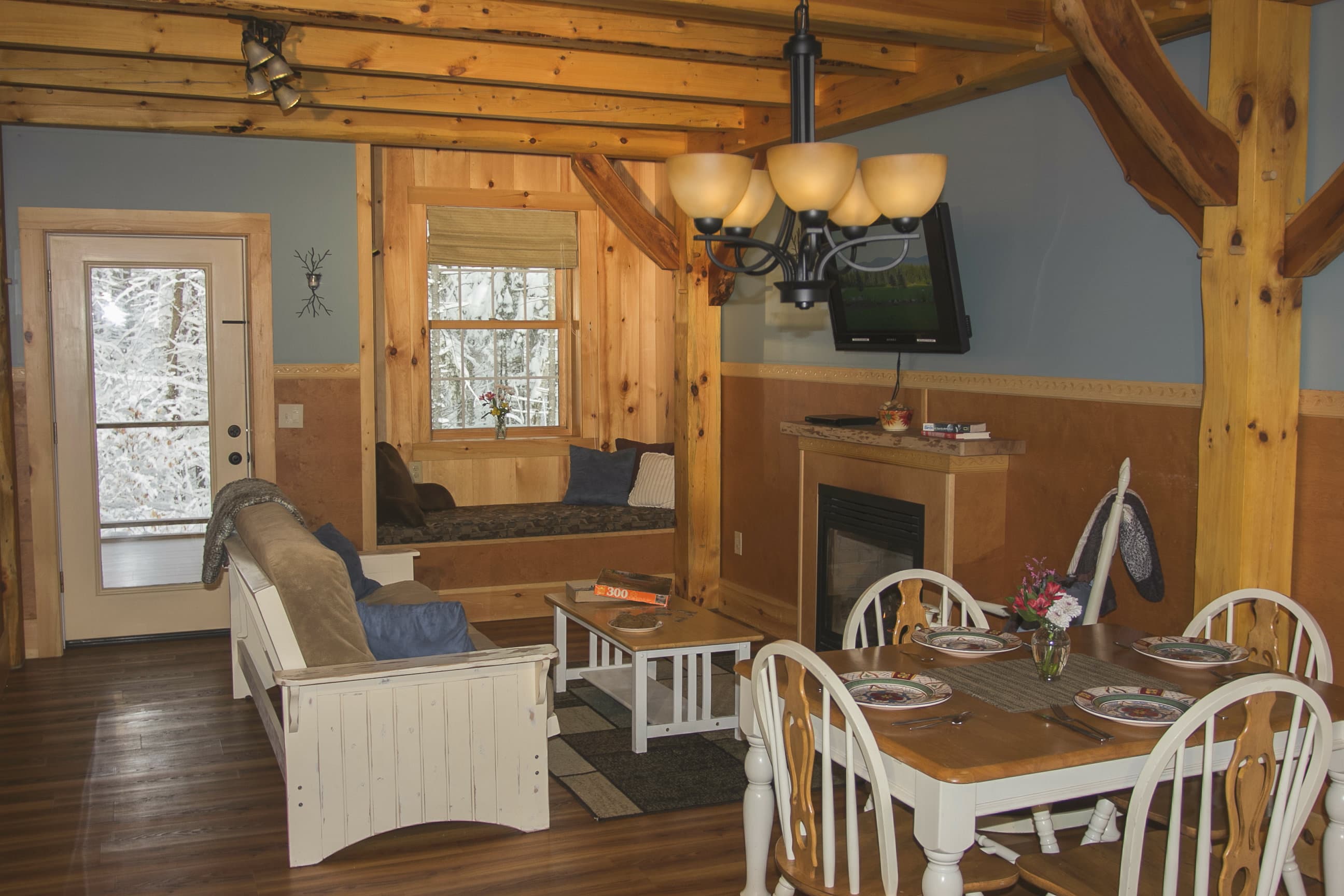 An open-concept living and dining area inside a rustic cabin features exposed wood ceiling beams and support posts. The space includes a white wicker sofa with blue pillows, a small wooden coffee table, and a square dining table with four white chairs. In the background, there is a built-in window seat, a small fireplace with a TV mounted above it, and a glass-paned door leading outside.
