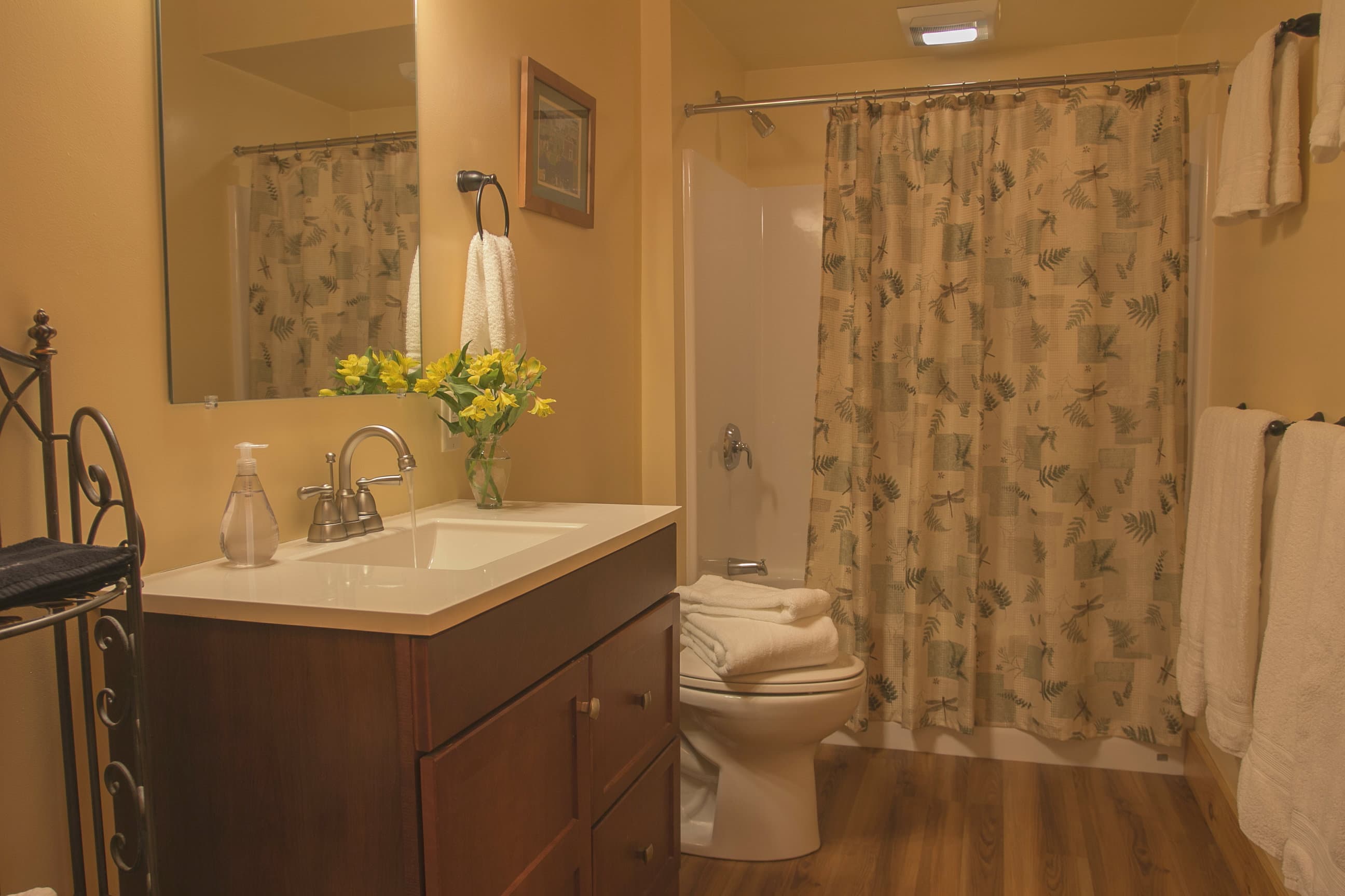 A warm, inviting cabin bathroom featuring a dark wood vanity with a white countertop and a large mirror reflecting the space. The room is decorated with yellow flowers in a vase, neatly folded white towels, and a floral patterned shower curtain that adds a touch of country charm to the neutral-toned walls.