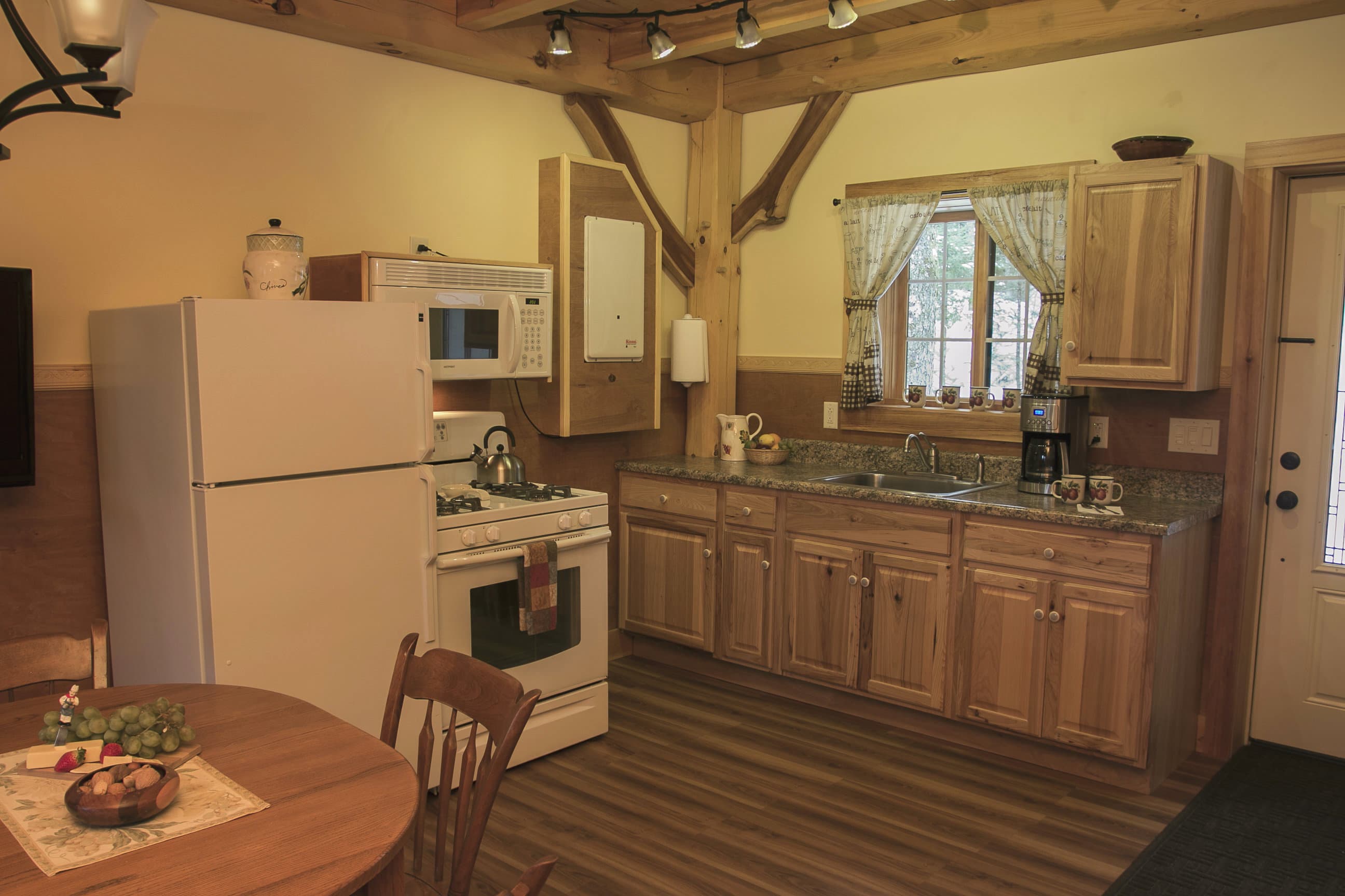 A warm, wood-paneled cabin kitchen and dining area featuring a light-colored round table set for a meal. The kitchen is equipped with light wood cabinetry, a white refrigerator, a gas stove, and a microwave, all accented by rustic timber-frame beams and a window with white café curtains.