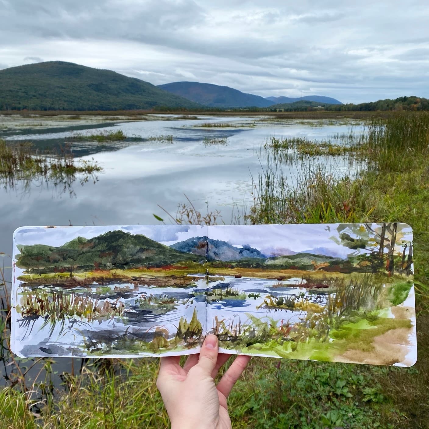 A person holds an open watercolor sketchbook depicting a tranquil landscape with mountains and water, set against a cloudy sky.
