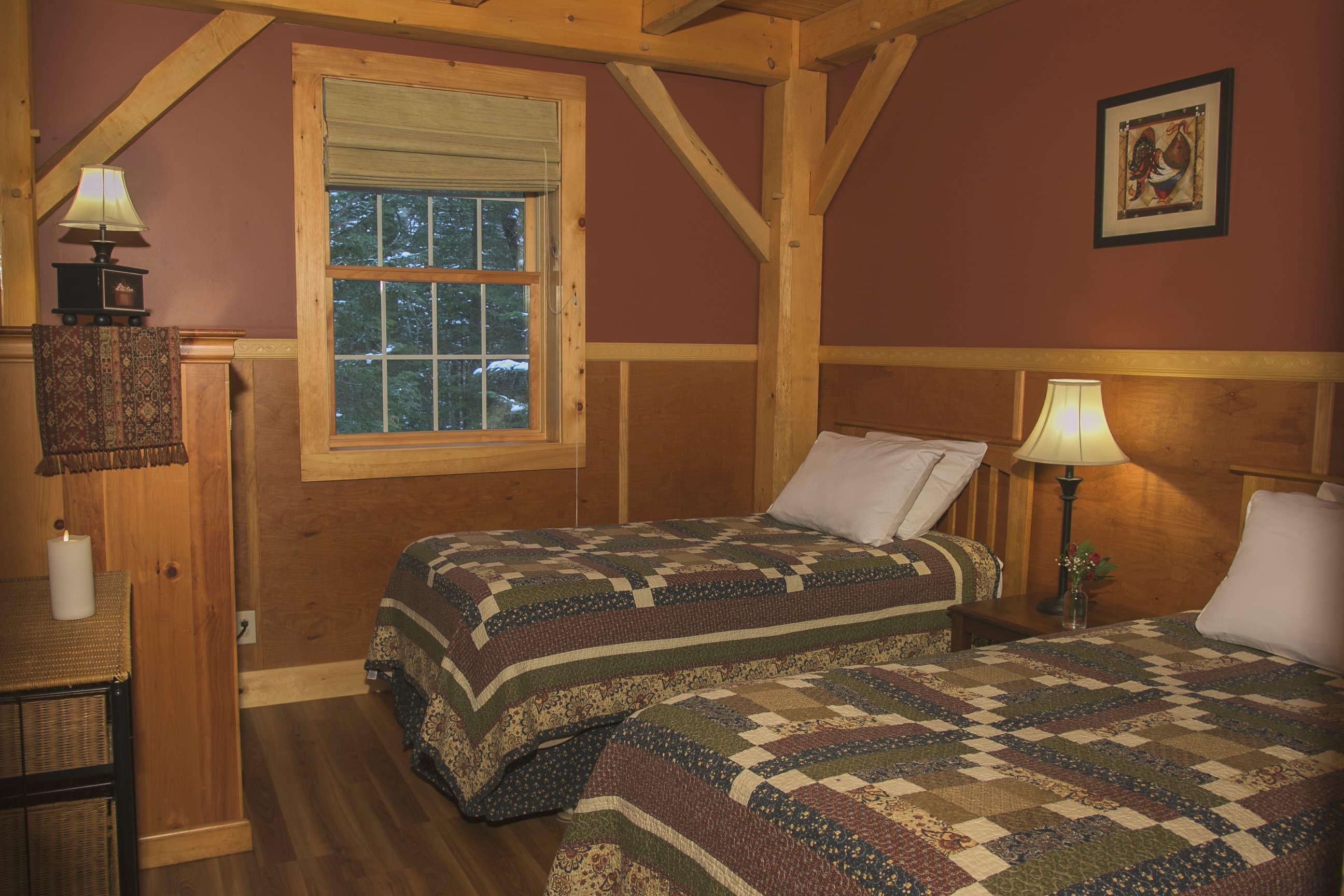 A cozy bedroom featuring two twin beds topped with green and brown patterned quilts, set against deep reddish-brown walls and rustic timber-frame beams. A window between the beds looks out onto a serene, wooded forest at Robert Frost Mountain Cabins.
