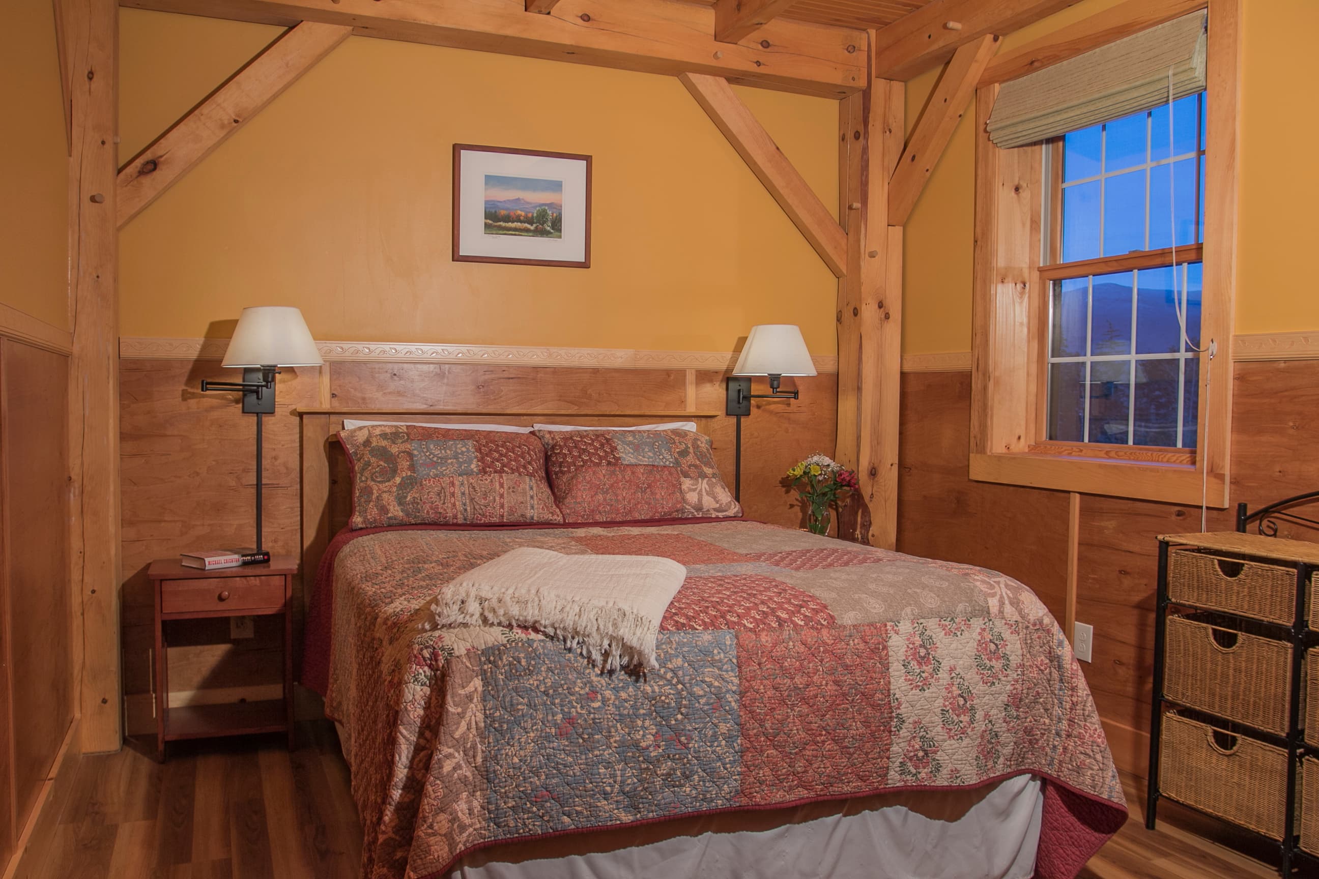 A cozy cabin bedroom featuring a queen-size bed with a patterned quilt, framed by rustic timber-beam architecture and warm, ochre-colored walls. The room includes bedside lamps, a small wooden nightstand, and a window offering a glimpse of the Vermont landscape.