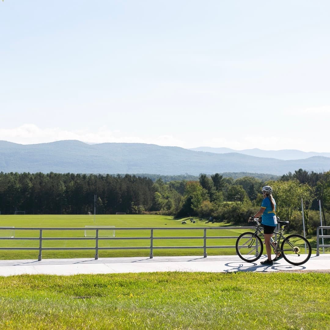A person in a helmet stands beside a bicycle, overlooking a green field and distant mountains under a clear blue sky.