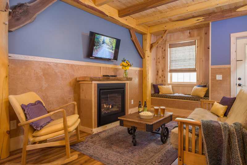A cozy living room inside a rustic cabin features a wooden armchair with a yellow cushion and purple pillow, a matching sofa, and a dark wood coffee table on a purple area rug. The space is decorated with periwinkle blue walls, wood wainscoting, and exposed timber beams, including a fireplace with a mounted TV and a built-in window seat in the background.