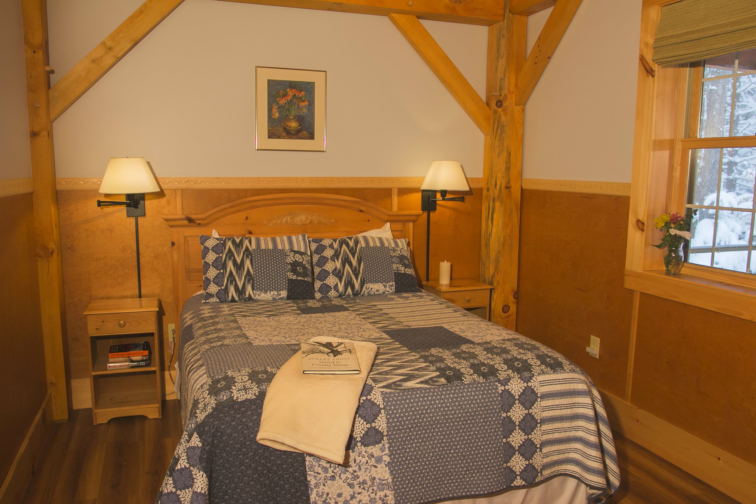 A cozy bedroom inside a rustic cabin features a bed with a blue and white patterned quilt, flanked by small wooden nightstands with matching lamps. Exposed timber beams frame the space, which has light-colored walls and a window revealing a snowy view outside.