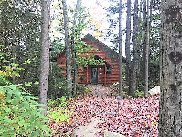 A cozy timber-frame cabin with reddish-brown wood siding, nestled deep within a lush green forest at Robert Frost Mountain Cabins in Ripton, Vermont. The cabin is surrounded by tall, slender trees, and the ground in the foreground is covered with fallen red and orange autumn leaves.