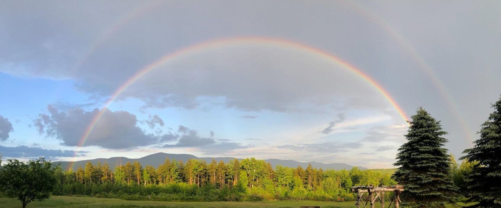 A double rainbow arches over lush green mountains and trees under a partly cloudy sky.