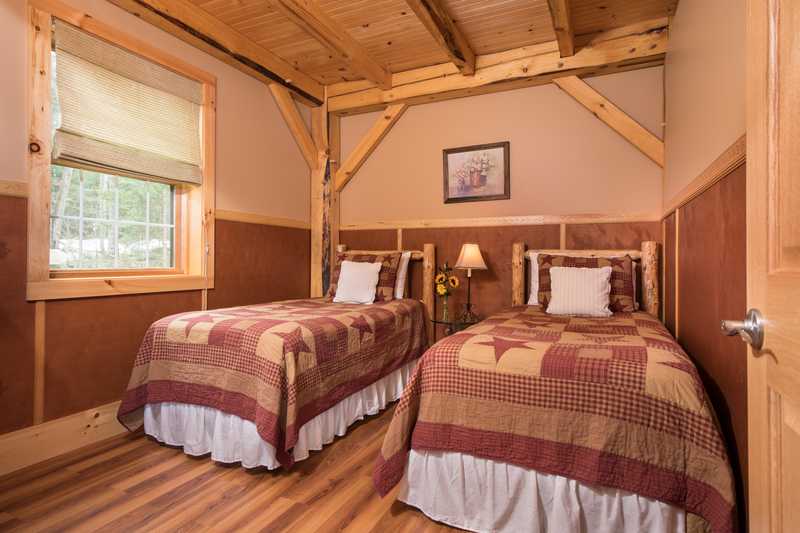 A bedroom inside a rustic cabin features two twin beds with log-style frames and warm, earth-toned patchwork quilts. The room is detailed with exposed timber framing, wood-paneled wainscoting, and wood-look flooring, with a window on the left wall and a small nightstand with a lamp centered between the beds.
