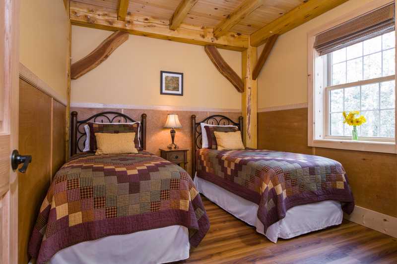 A bedroom inside a rustic cabin features two twin beds with dark metal frames and earth-toned patchwork quilts. The room is designed with light tan walls, warm wood wainscoting, and exposed timber framing, with a window on the right wall and a single nightstand with a lamp positioned between the beds.