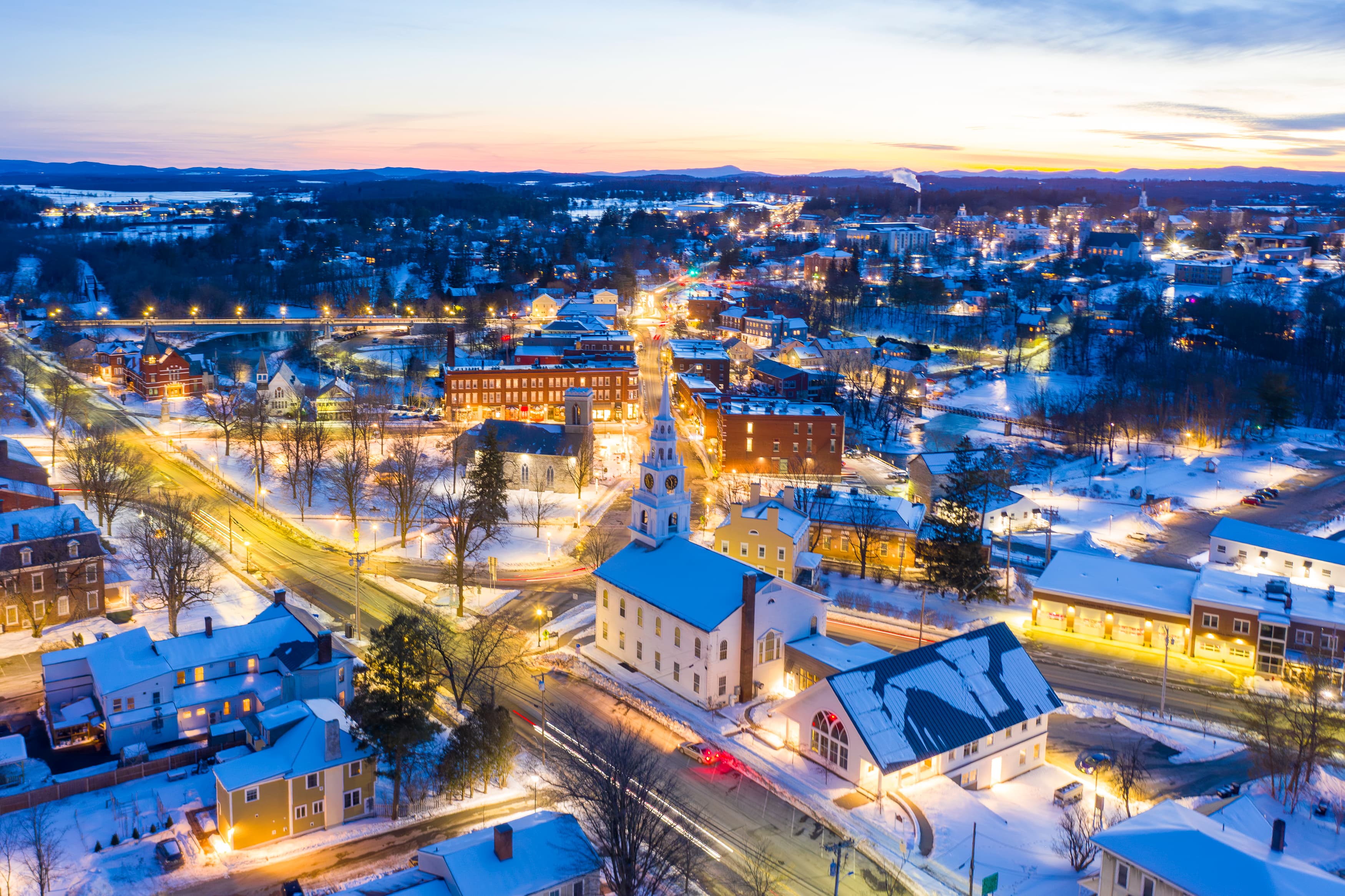 Aerial view of a snow-covered town at dusk, illuminated by streetlights and featuring a prominent church.