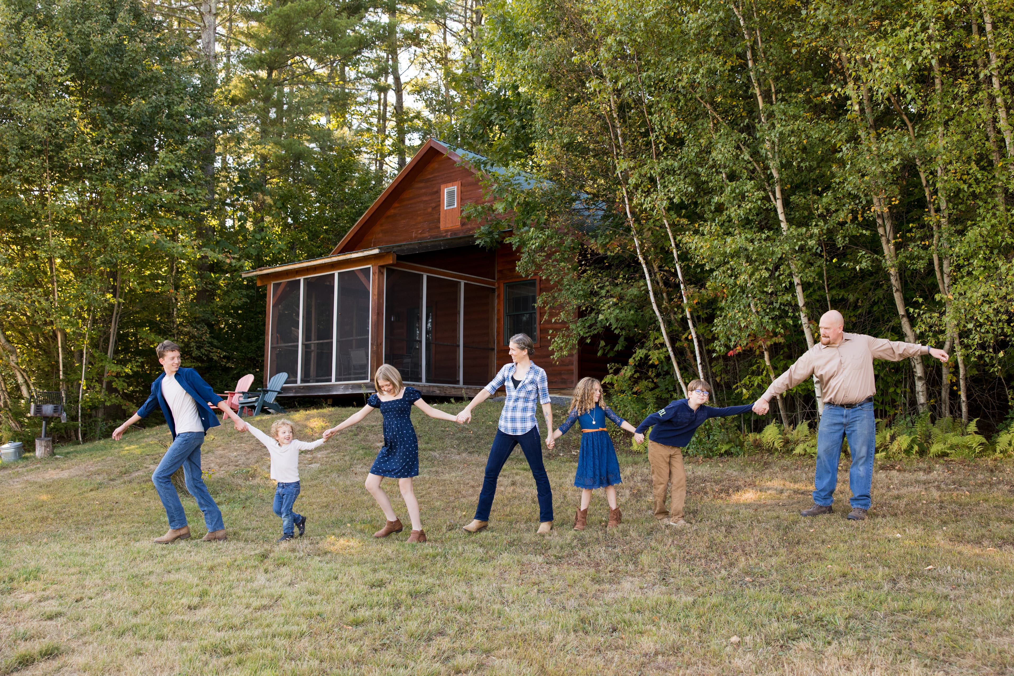 A family holding hands and smiling in front of a cabin amidst trees.