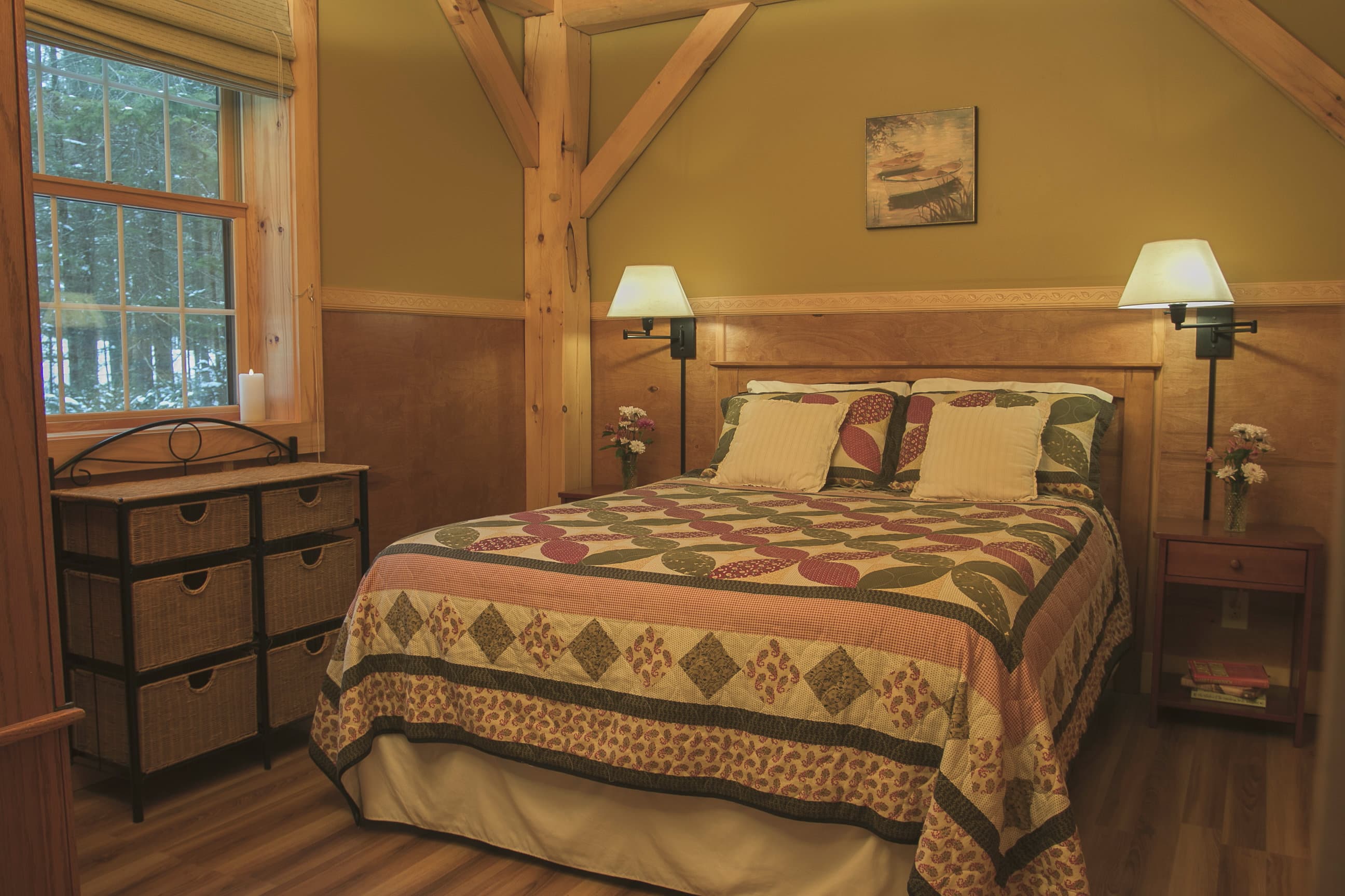 A cozy cabin bedroom featuring a queen-size bed with a large, colorful diamond-patterned quilt. The room is defined by rustic timber-frame beams, warm olive-toned walls, and wood-paneled wainscoting. A large window on the left reveals a view of the wooded forest outside, and the space is finished with a wooden dresser and matching bedside nightstands with lamps.