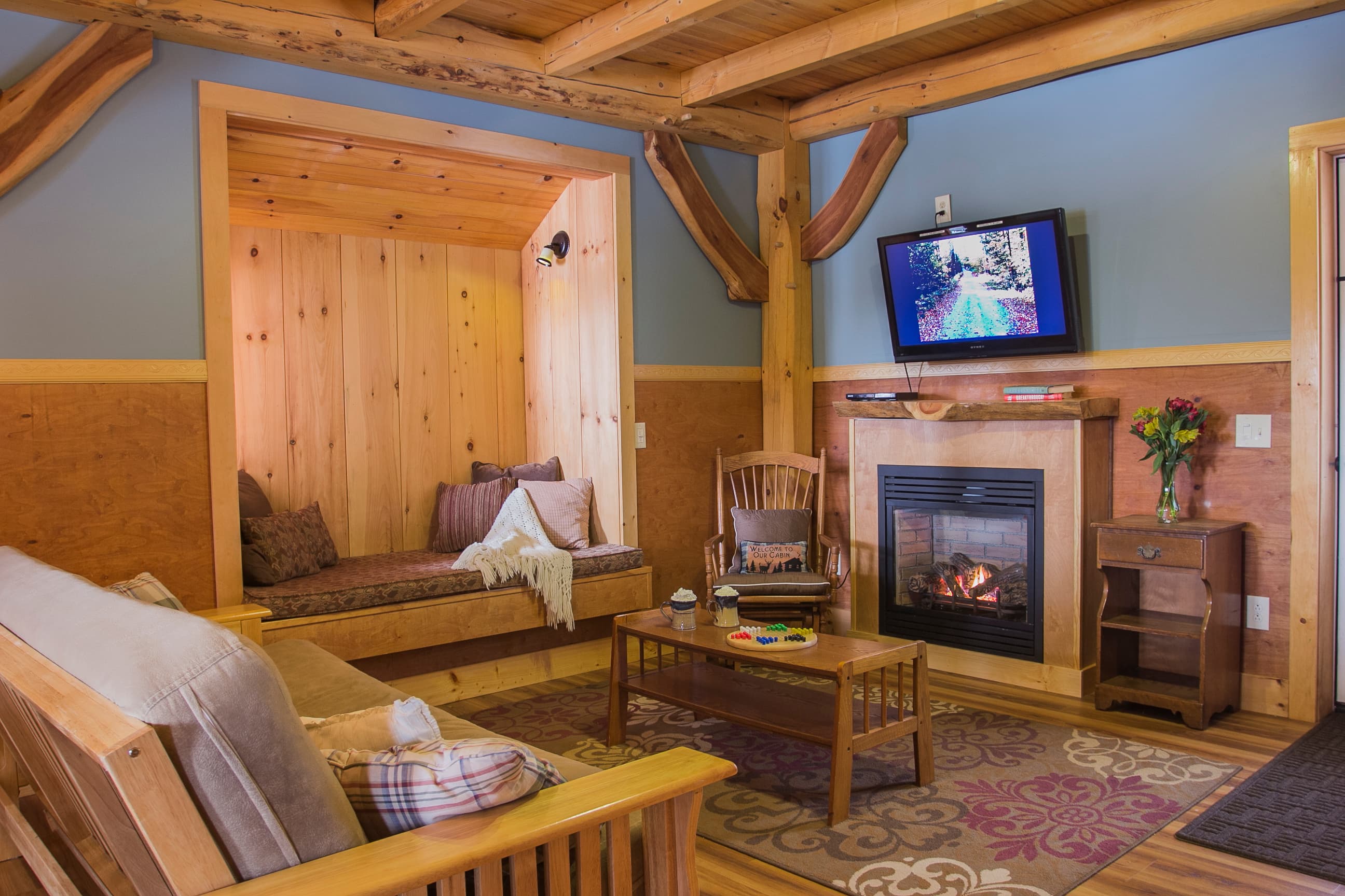 A cozy cabin living room featuring exposed timber beams, a glowing fireplace, and a unique wood-lined Swedish reading nook tucked into the wall. A comfortable futon and coffee table sit on a patterned rug, creating a warm and inviting mountain retreat.