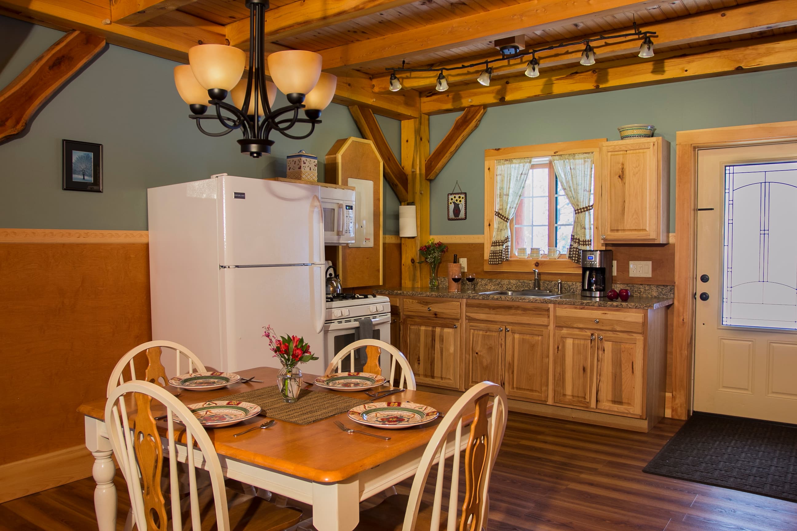 The interior of a rustic cabin features a kitchen and dining area with warm wood accents and exposed timber framing. A wooden dining table with four white chairs is set in the foreground, illuminated by a classic chandelier. The kitchen in the background includes light wood cabinetry, a white refrigerator, a white stove, and a window with light curtains.