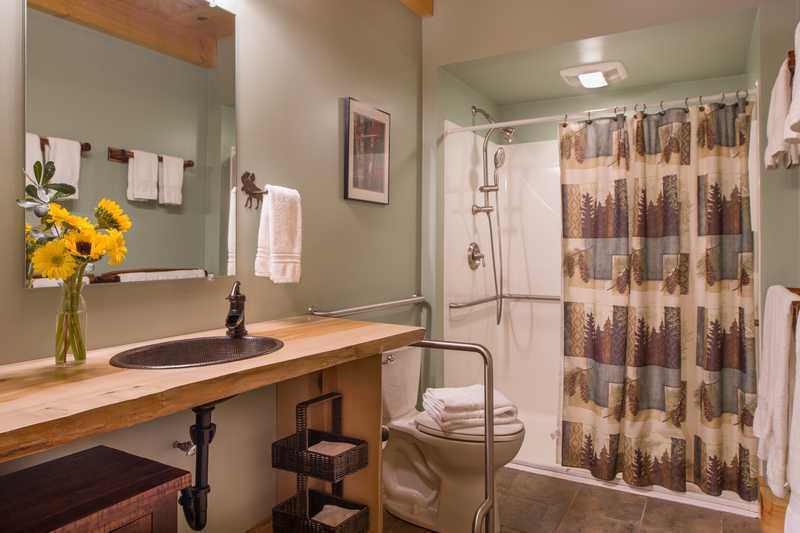 An accessible bathroom in a rustic cabin features an open wood-slat vanity with a black drop-in sink and a large wall mirror. The space includes a toilet with metal grab bars, a walk-in shower with a tree-patterned curtain, sage green walls, and warm wood flooring.