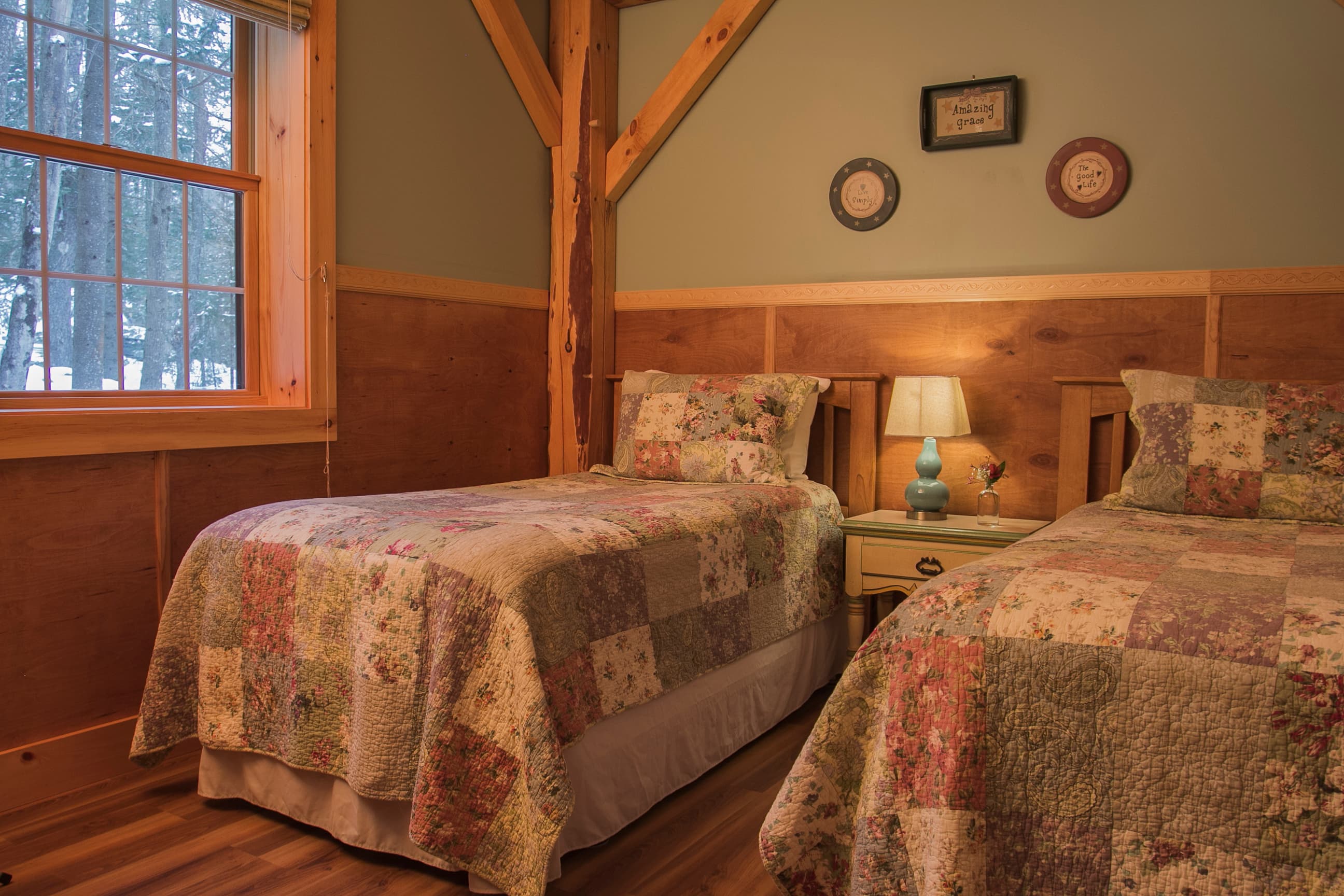 A cozy cabin bedroom featuring two twin beds with floral patchwork quilts, separated by a small nightstand with a turquoise lamp. The room showcases rustic timber-frame architecture, warm wood paneling, and a large window overlooking a peaceful, wooded forest.