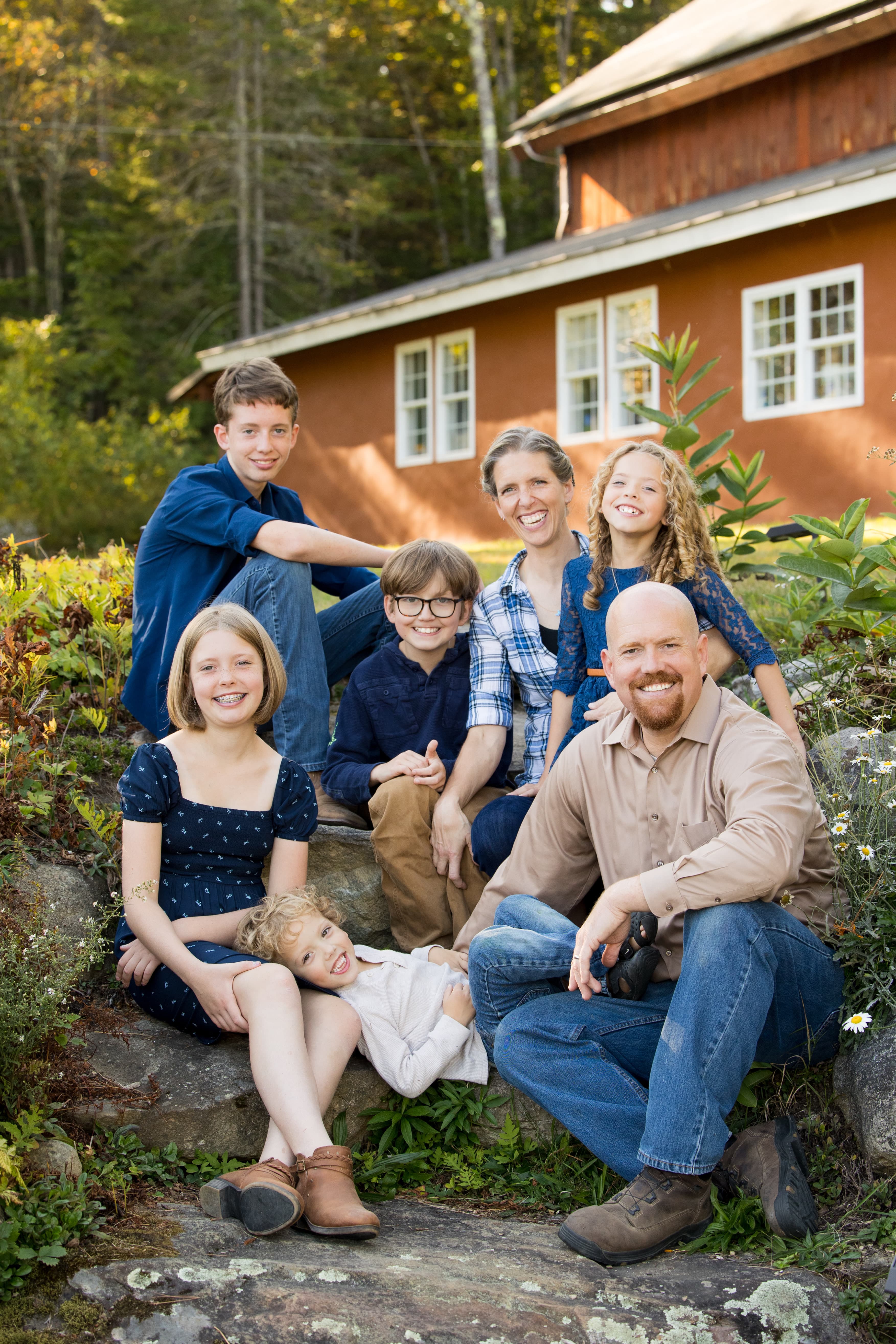 A family of six sits together on a stone, smiling in front of a rustic house and lush greenery.