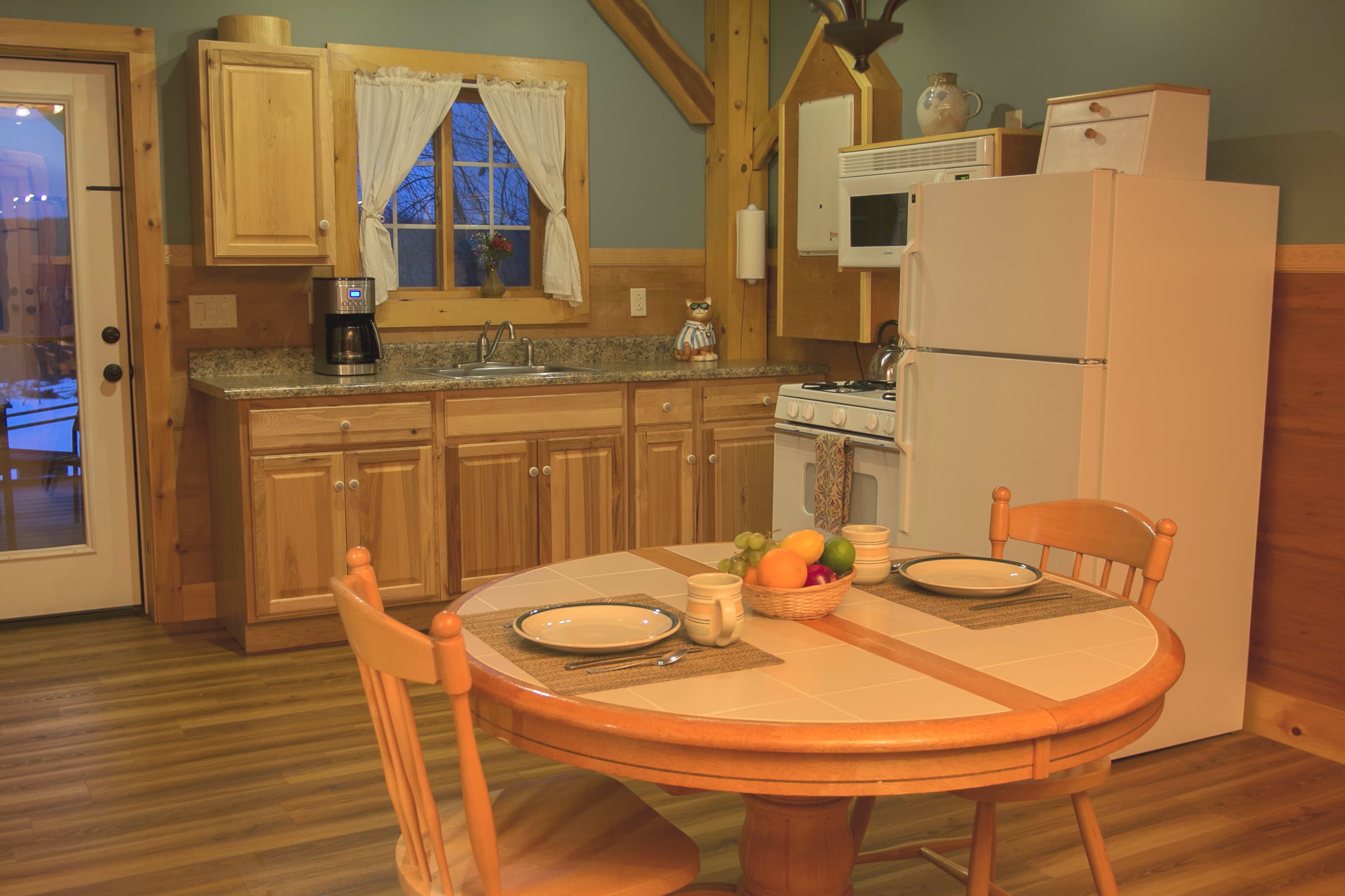 An inviting cabin kitchen and dining area featuring a round wooden table set for two. The space includes natural wood cabinetry, a full-sized white refrigerator, and a window with white curtains, all framed by rustic timber-frame beams.