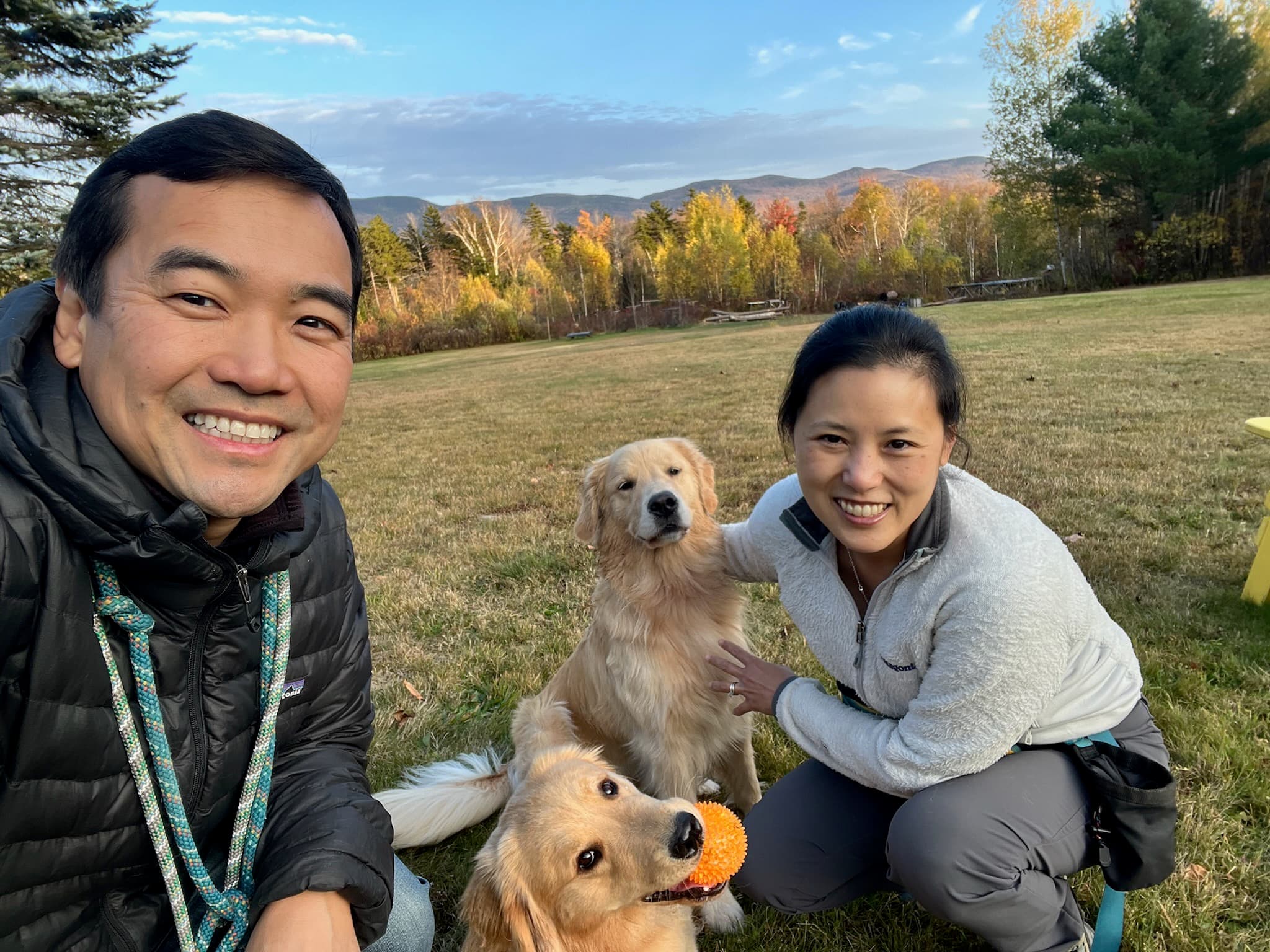 A man and a woman smile for a selfie with two golden retrievers in a grassy field with trees in the background.