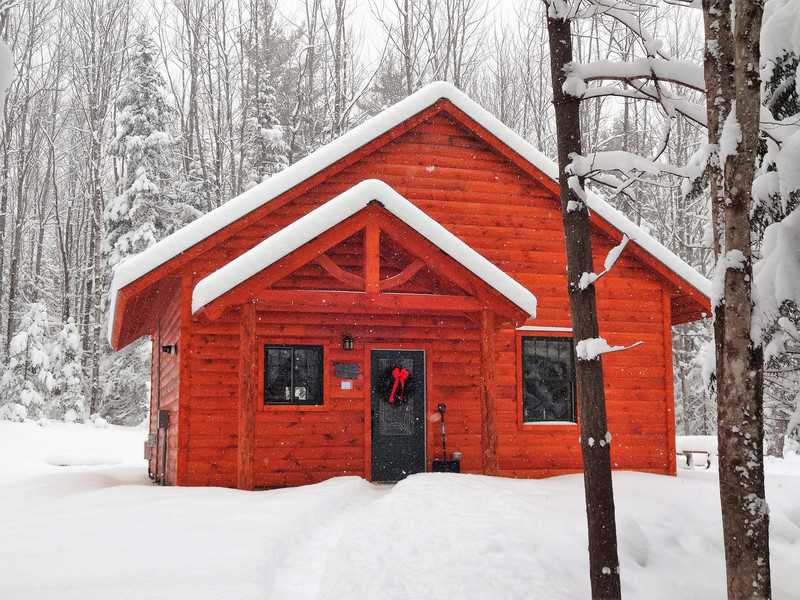 A small, wood-sided cabin with a peaked roof and a timber-frame porch entryway nestled in a snow-covered forest. The cabin's bright orange-toned wood creates a warm contrast against the white snow on the ground and the frosted trees under a clear winter sky.