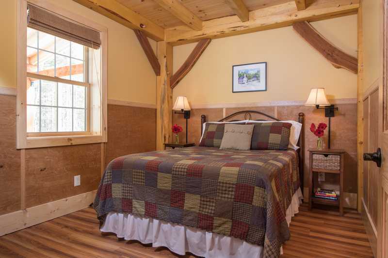 A bedroom in a rustic cabin featuring a bed with a dark metal frame and an earth-toned patchwork quilt. The room is designed with light tan walls, warm wood wainscoting, and exposed timber ceiling beams. A window with a neutral-colored shade is on the left wall, and two small bedside tables with lamps flank the bed.