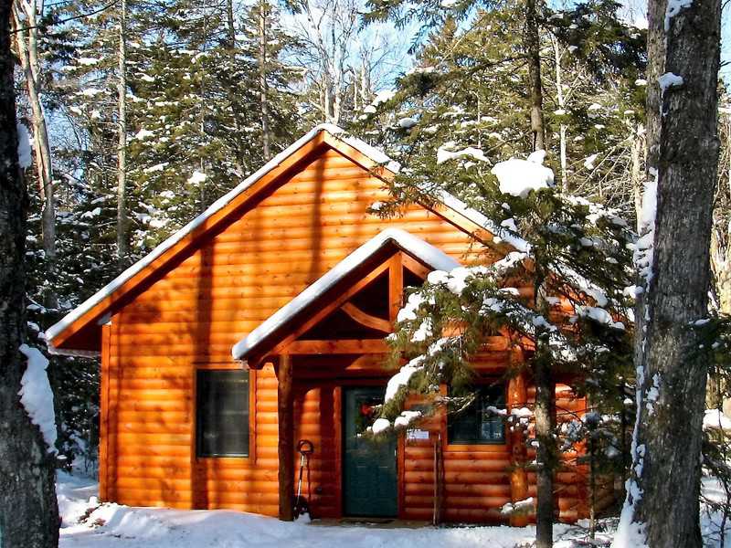 A small, rustic wood-sided cabin with a peaked roof and a timber-frame porch entryway sits nestled in a snowy evergreen forest.