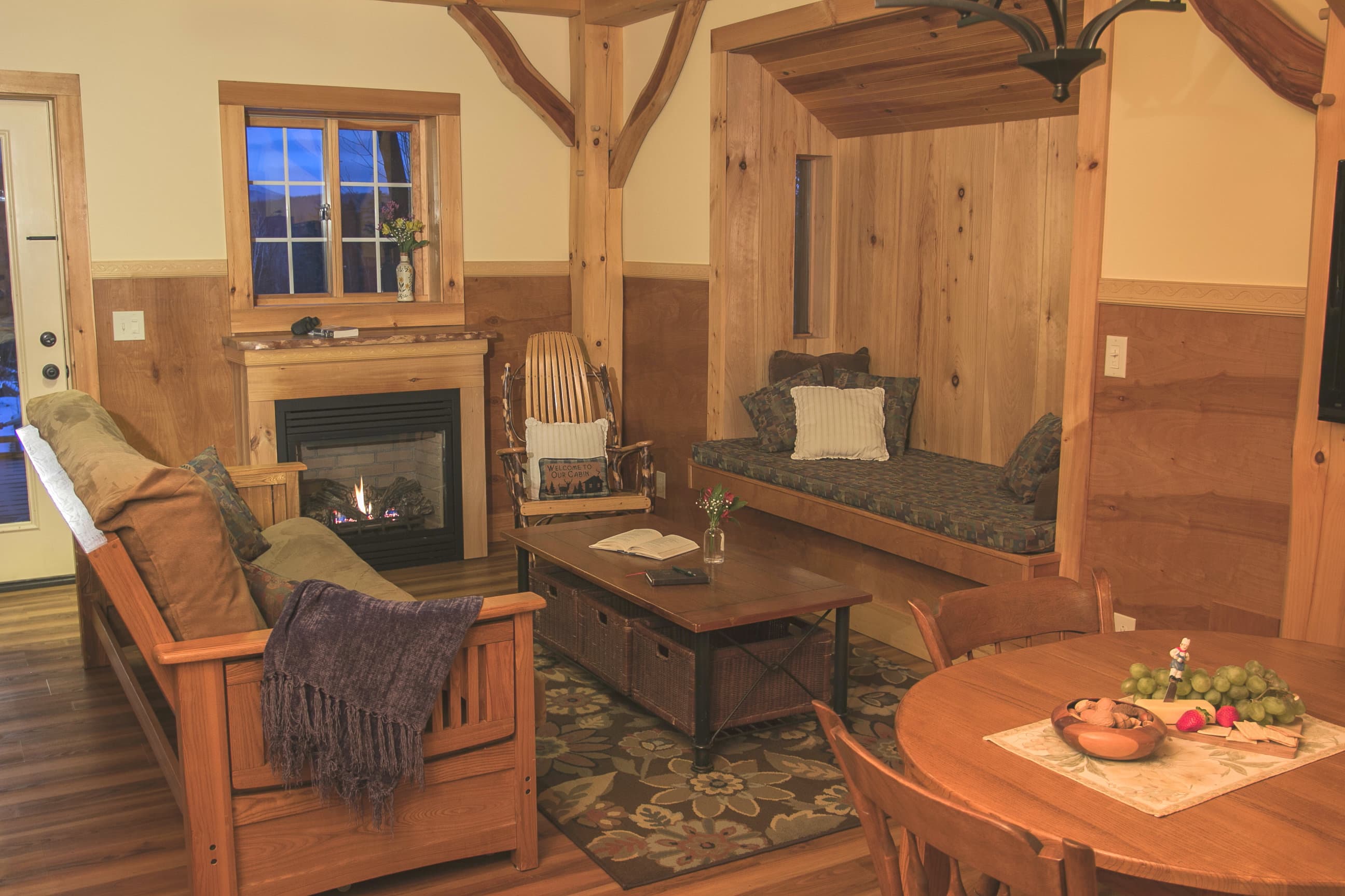 A warm and cozy cabin living area featuring a fireplace, a comfortable futon, and an artisan-crafted Swedish reading nook built into the timber-frame wall. The room is decorated with rustic wooden furniture, a floral rug, and seasonal flowers, embodying the quiet mountain retreat atmosphere of Robert Frost Mountain Cabins.