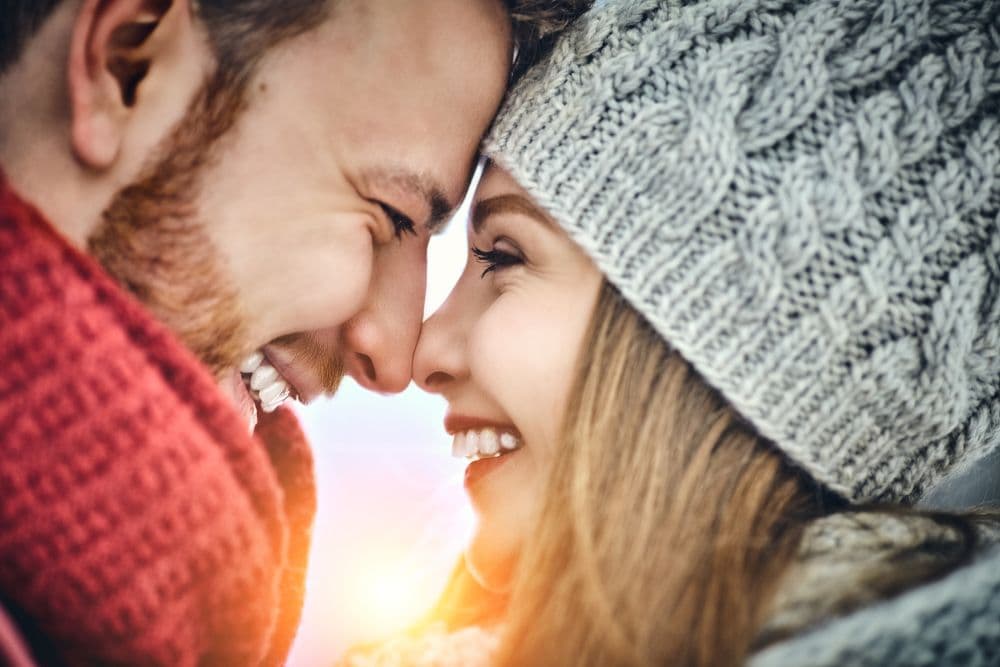 A joyful couple smiles at each other while gently touching foreheads, wearing winter scarves and hats.