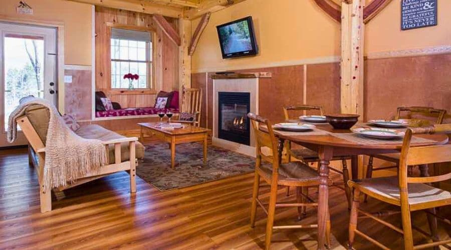 A cozy living and dining space featuring a fireplace, wooden furniture, and large windows.
