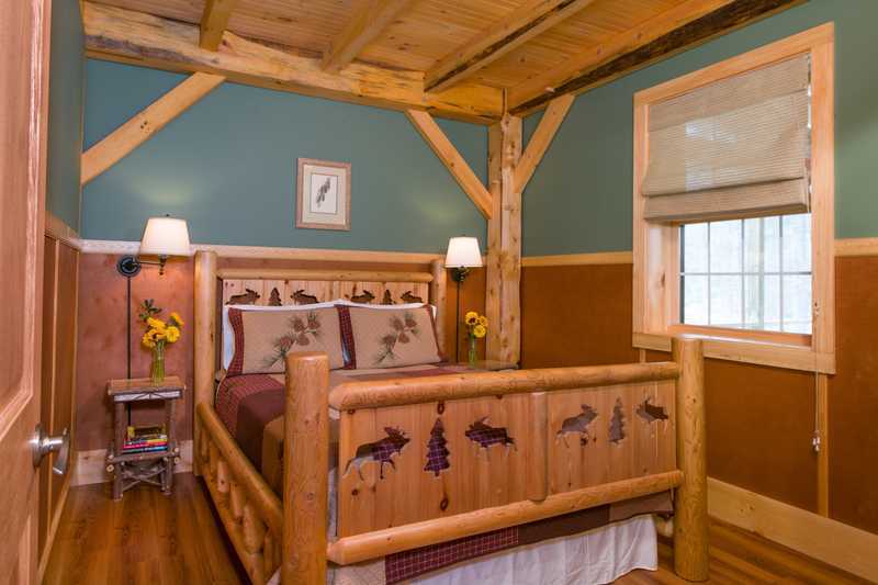 A bedroom inside a rustic cabin features a bed with a log-style frame and wildlife-themed bedding. The room is decorated with teal walls, warm wood wainscoting, and exposed timber ceiling beams, with a window on the right wall and small lamps on the nightstands.