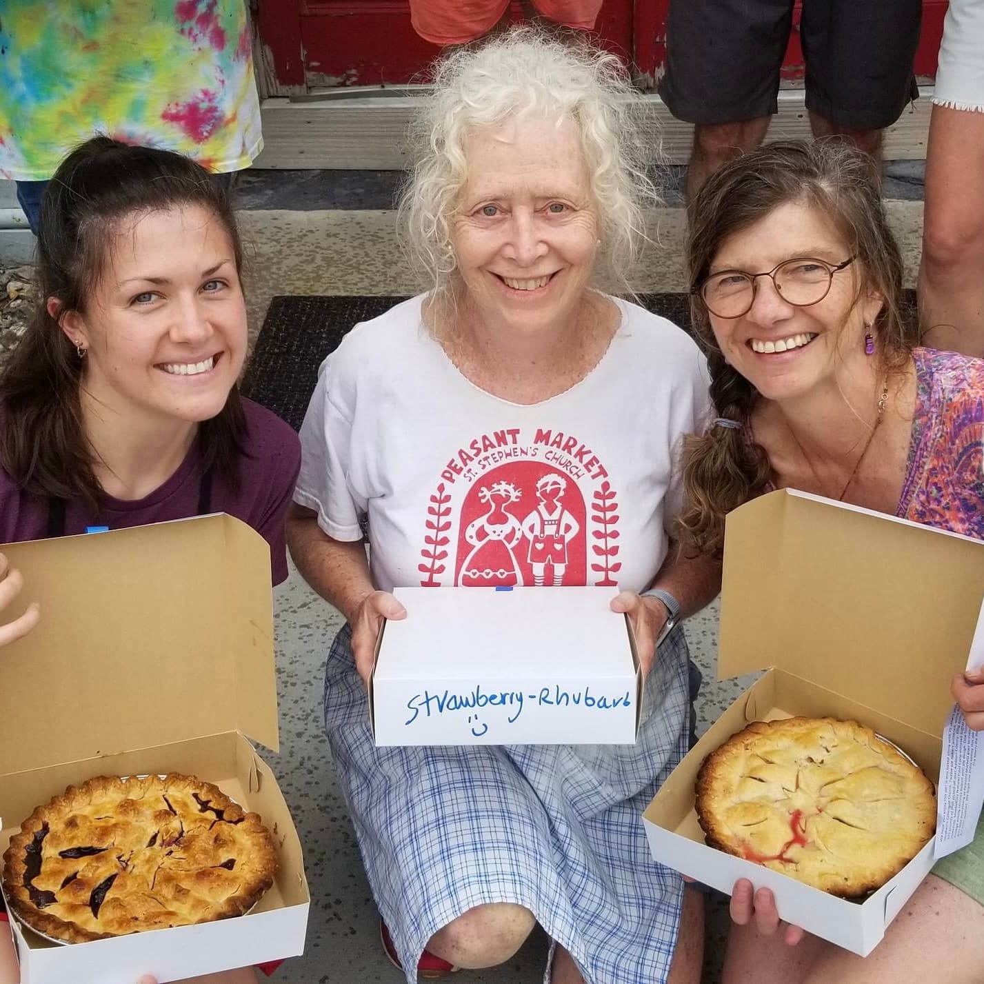 Three women smile while holding boxes containing pies, with one pie labeled "Strawberry-Rhubarb."