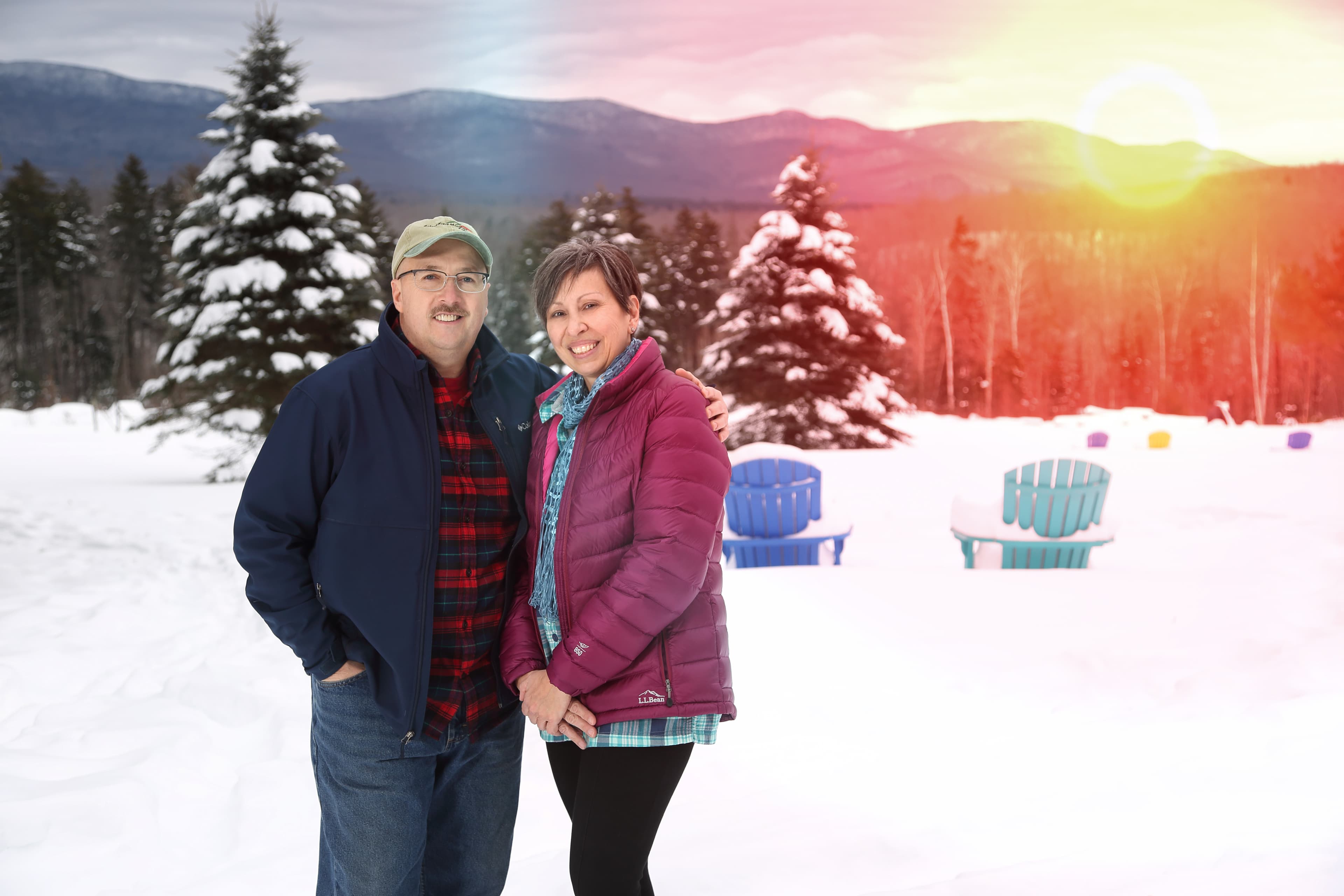 A couple stands together in a snowy landscape with mountains and colorful chairs in the background.