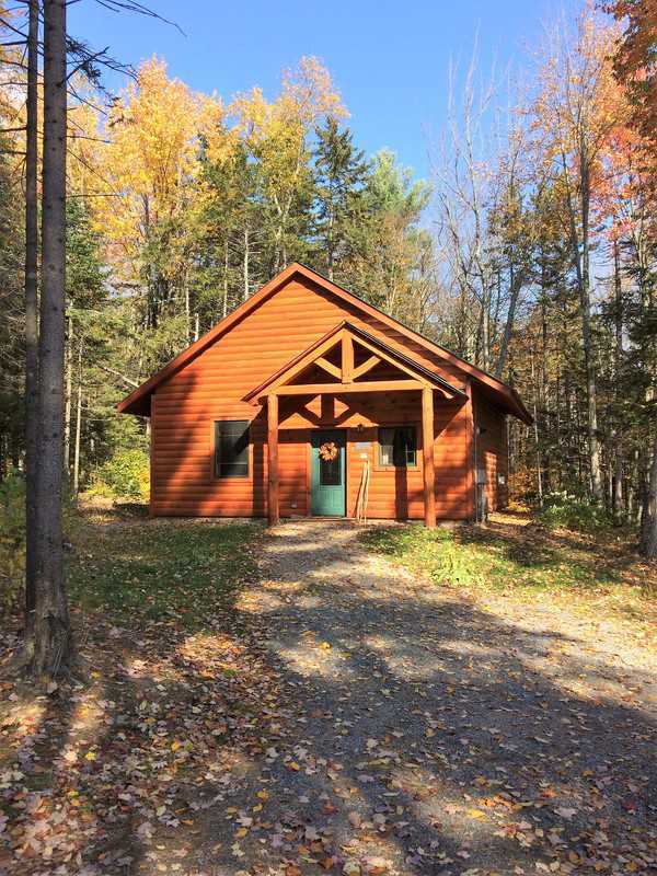 A small, rustic wood-sided cabin with a peaked roof and a timber-frame porch entryway sits nestled in a forest of tall, thin trees with yellow autumn leaves under a clear blue sky.