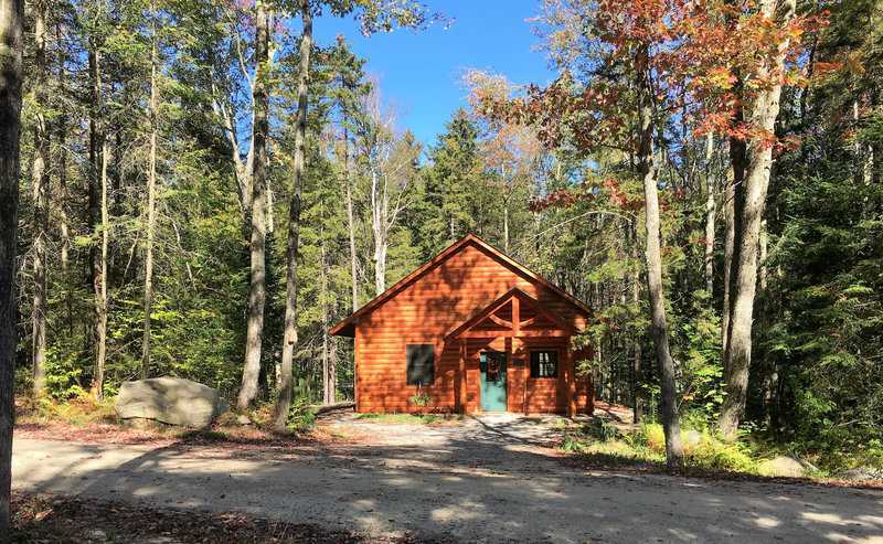 A small, rustic wood-sided cabin with a peaked roof and a timber-frame porch entryway, set back from a gravel driveway in a lush green forest with hints of autumn yellow under a clear blue sky.