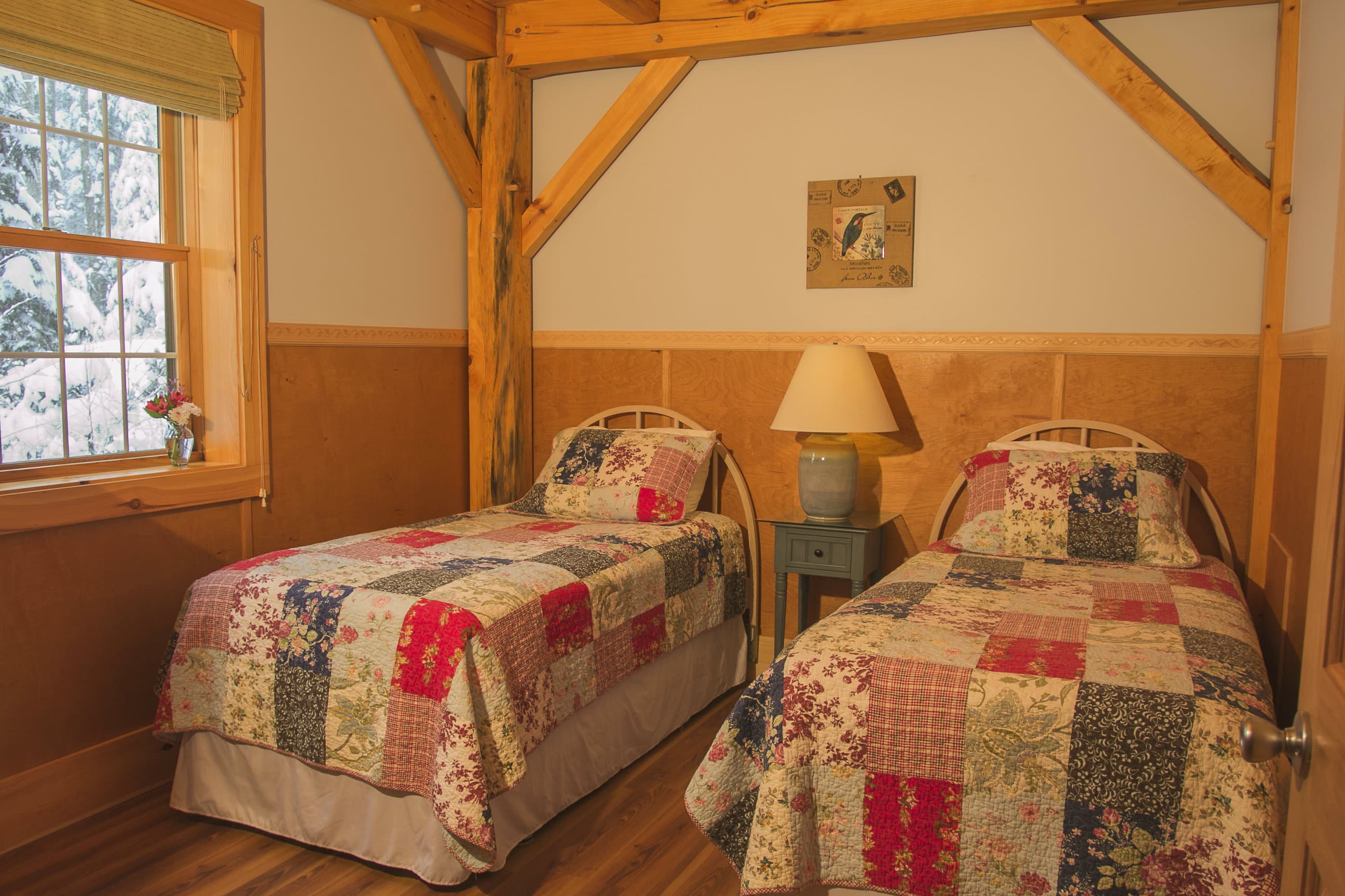 A bedroom inside a rustic cabin contains two twin beds with colorful, multi-patterned patchwork quilts. A single wooden nightstand with a lamp sits between the beds, and the room features exposed timber framing, warm wood-paneled lower walls, and a window showing a snowy landscape.